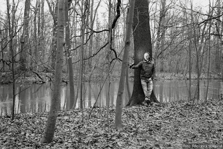 Mann steht im Wald vor einem Baum mit einem Blitzlicht in der Hand, welches ihn anleuchtet.